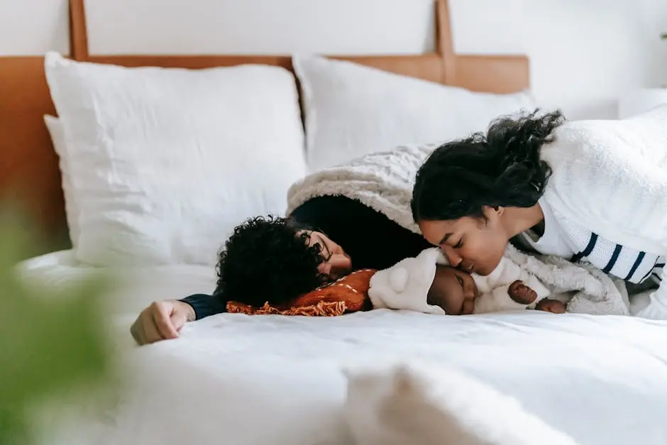 Happy young African American mother kissing adorable baby sleeping on cozy bed with ethnic father in sunny morning