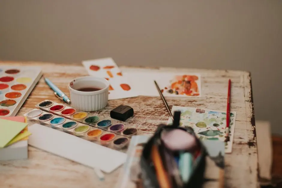 A cluttered artist’s desk with watercolor paints, brushes, papers, and a cup, offering a glimpse into creative processes.