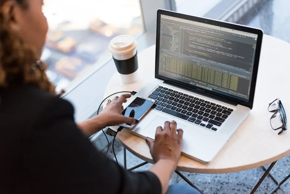 Black woman programming on a laptop with coffee, smartphone, and glasses on a desk in an office.