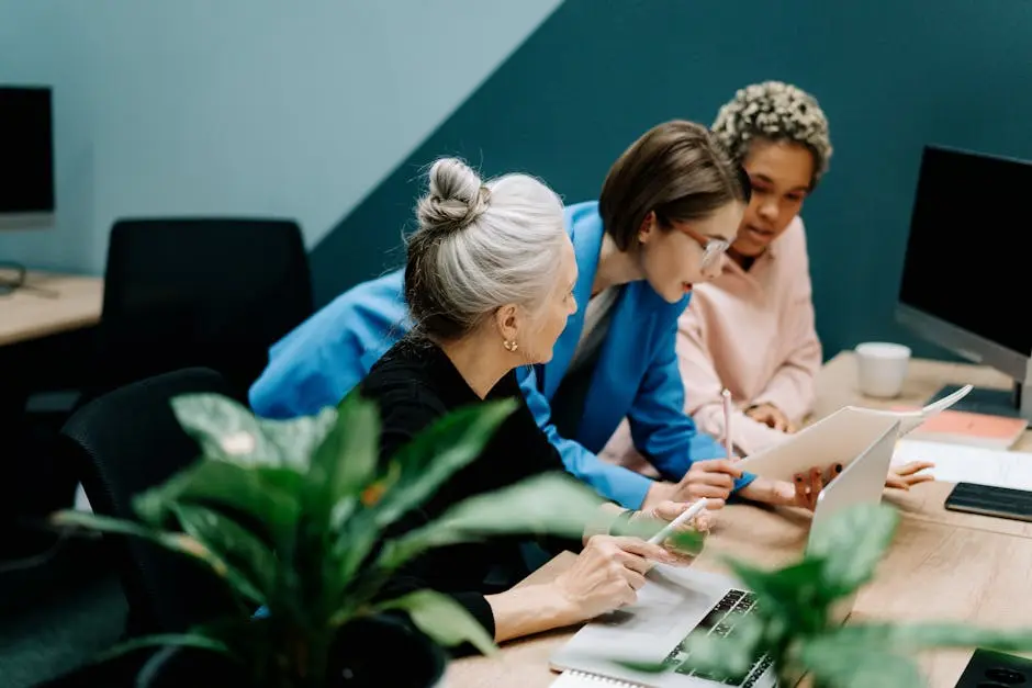 Three professionals collaborating in a modern office setting, discussing internal communications strategies with laptops and documents, surrounded by greenery.