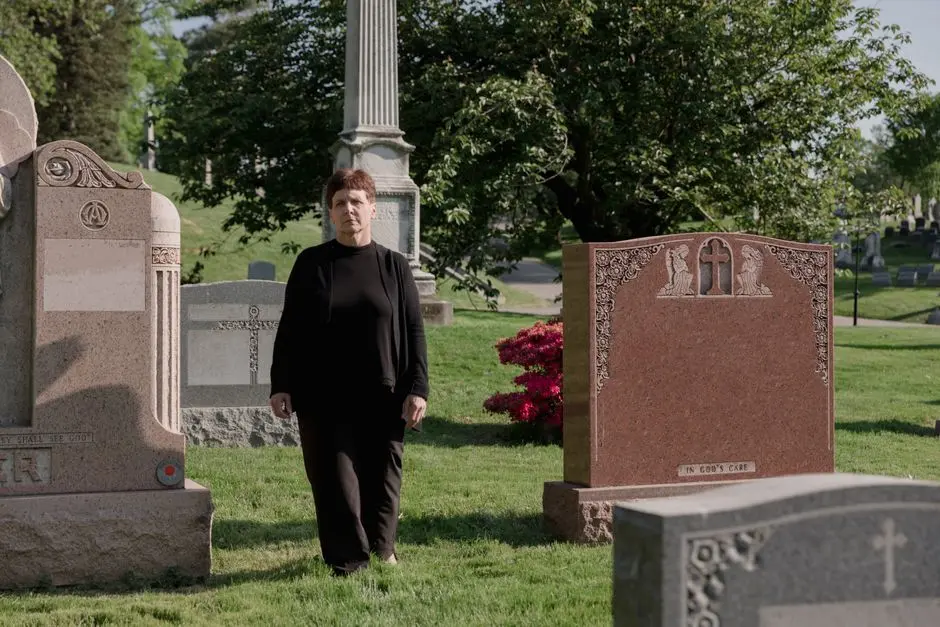 A woman in black clothes stands solemnly among gravestones in a serene cemetery.