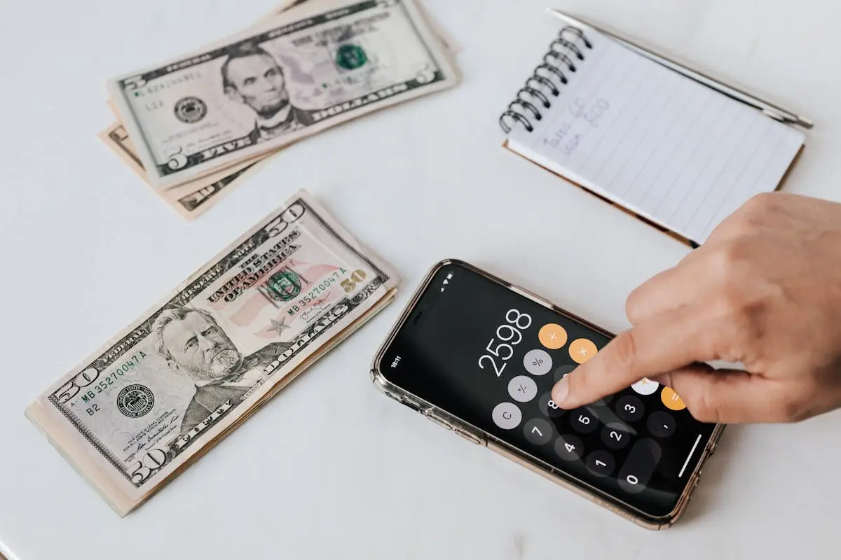 From above of crop anonymous economist calculating total amount of income using calculator app on cellphone near piles of greenbacks and notebook with pen, Interim CFO Services