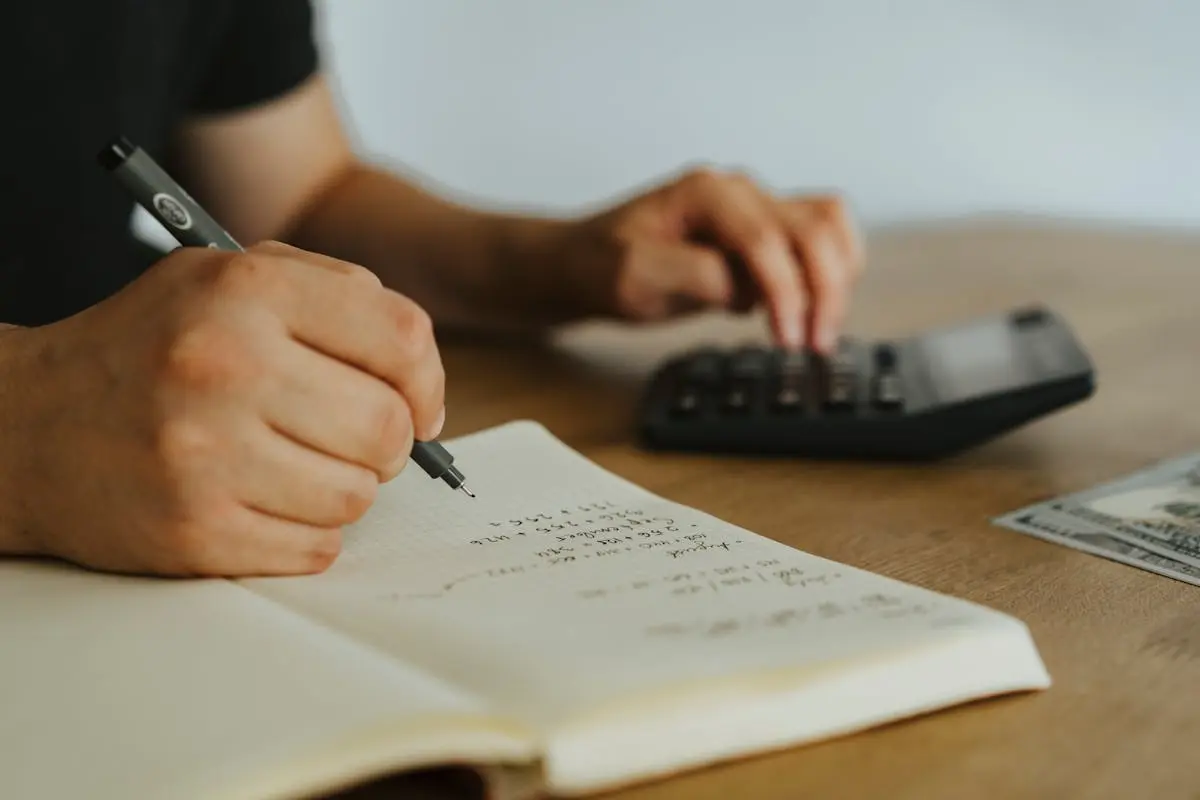 Close-up of hands writing calculations in a notebook with a calculator, focused on budgeting or business acquisitions. 