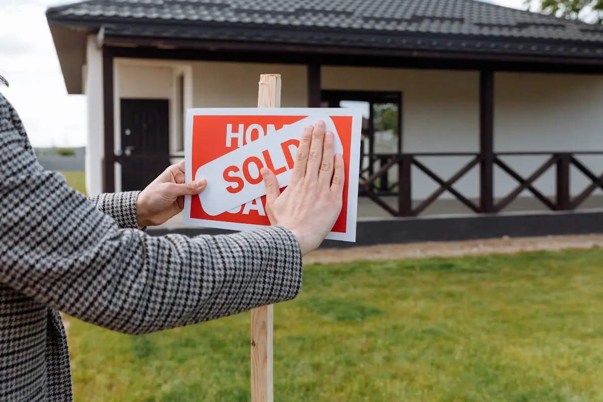 Close-up of hands placing ‘sold’ sticker on sign in front of a new house.