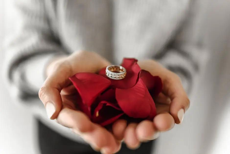 Close-up of an engagement ring resting on red rose petals held in hands, symbolizing love and commitment.