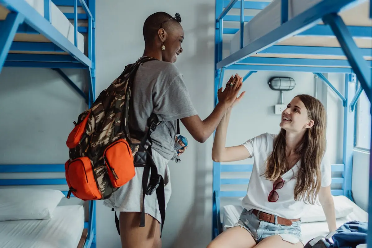 Two female backpackers high-fiving in a bright hostel dorm room, enjoying travel adventures.