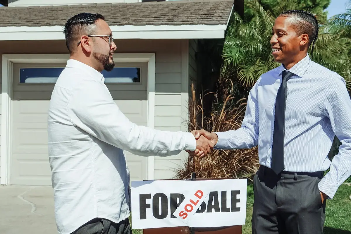 Two men shaking hands in front of house sold sign, sealing real estate deal outdoors.