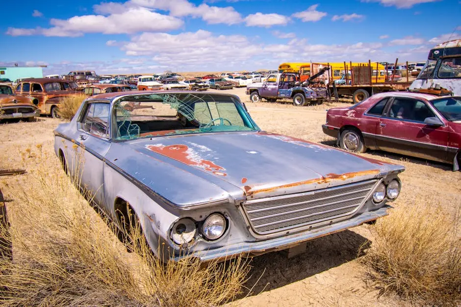 A rusty old car sits amidst other vintage vehicles in an open desert junkyard.