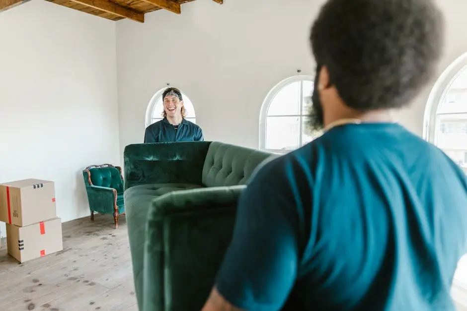 Two movers carrying a green couch in a sunlit room with packed boxes.