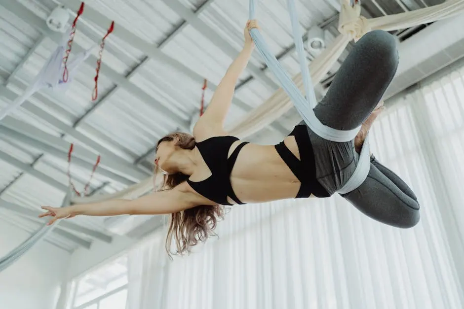 Young woman in sportswear performing aerial acrobatics indoors suspended by fabric.