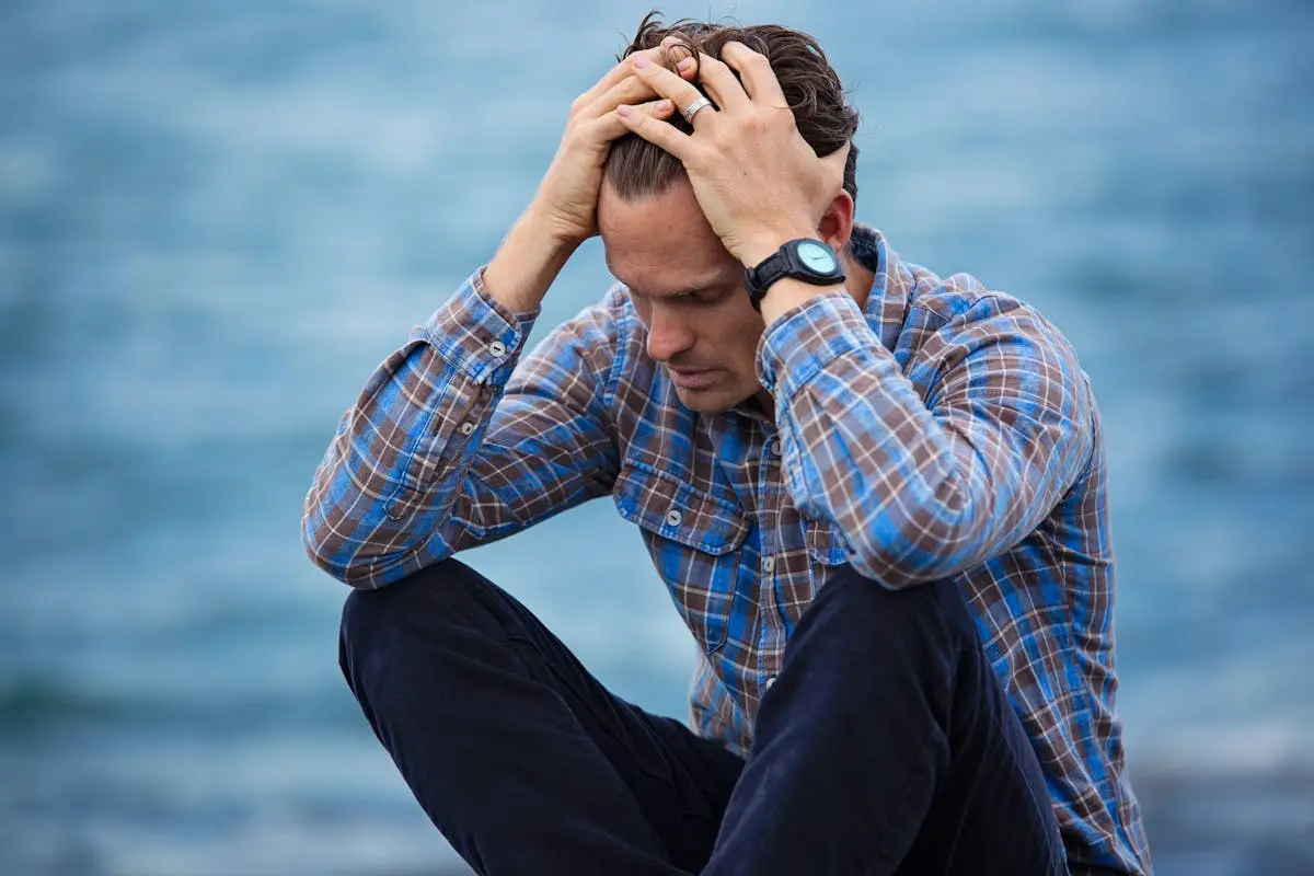 A man in a plaid shirt sits by the water looking distressed, symbolizing stress and thinking about Career Change.