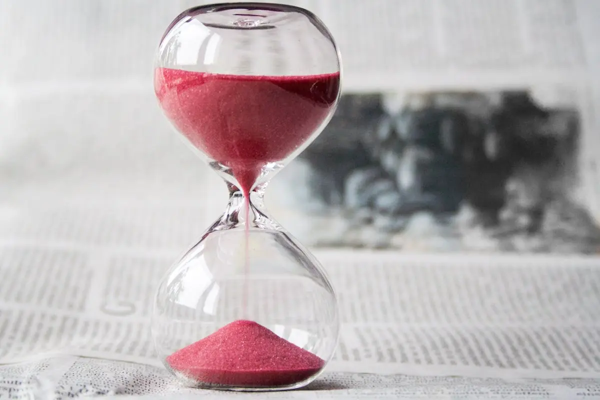 Close-up of a transparent hourglass with pink sand flowing, placed on a newspaper background.