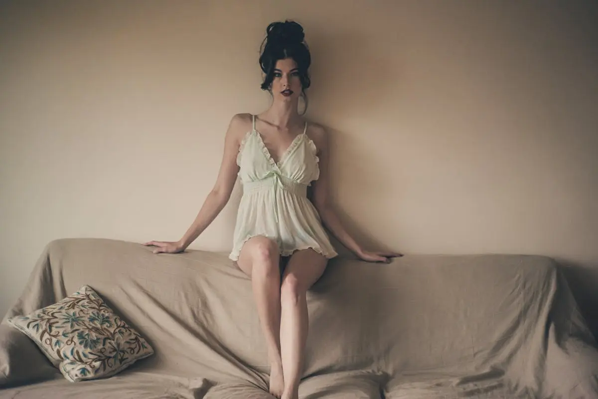 A brunette woman in lingerie posing gracefully on a sofa in a soft, moody indoor setting.