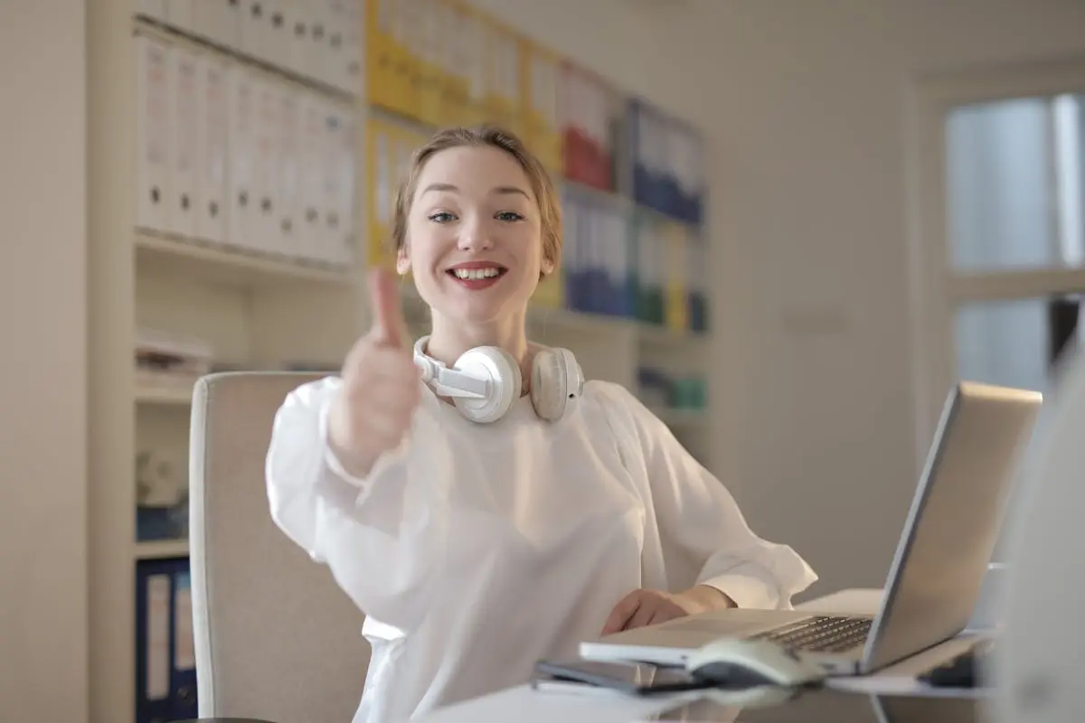 Smiling woman giving thumbs up while working with laptop and headphones in an office.