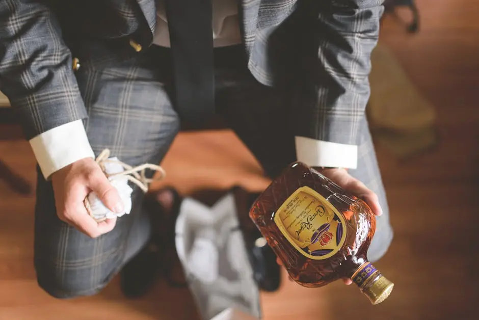 A stylish man in a plaid suit holding a whiskey bottle and a small cloth bag indoors.