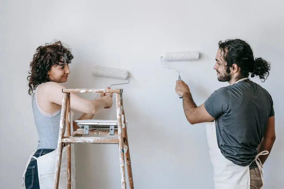 Cheerful adult ethnic couple in aprons looking at each other while painting wall with rollers during renovation process in house