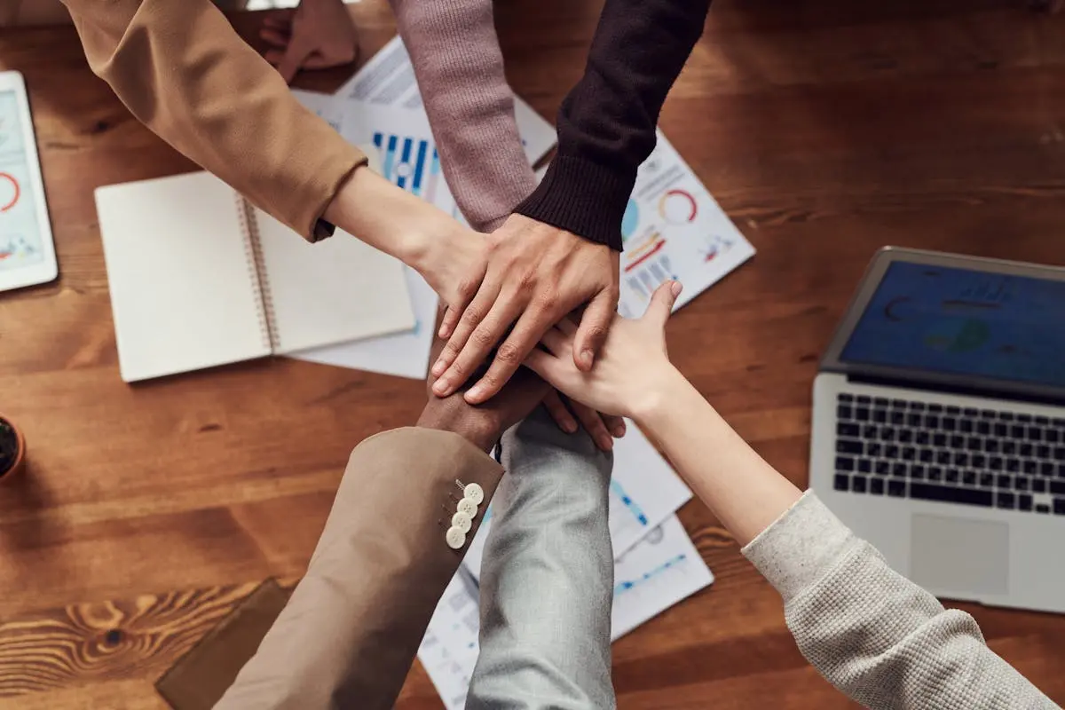 Diverse business coach professionals unite for teamwork around a wooden table with laptops and documents.