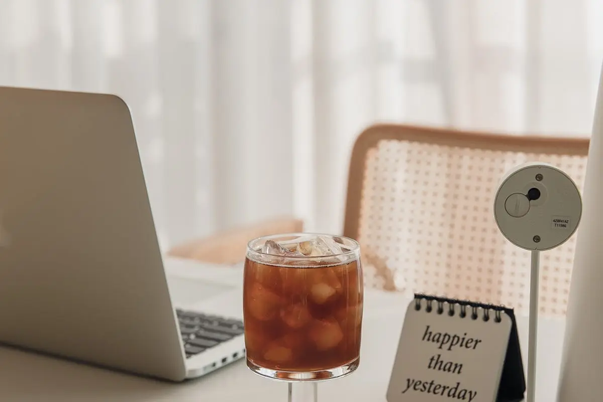 Cozy workspace featuring a laptop, iced coffee, and motivational note in soft lighting.