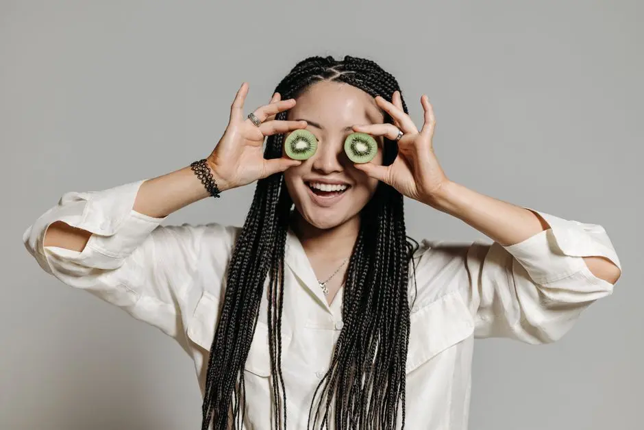 Woman with braided hair playfully holding kiwi slices over her eyes, smiling in a casual setting.