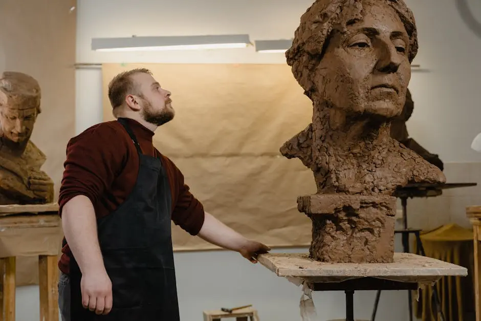 A male sculptor wearing an apron works on a detailed clay bust sculpture in an art studio.