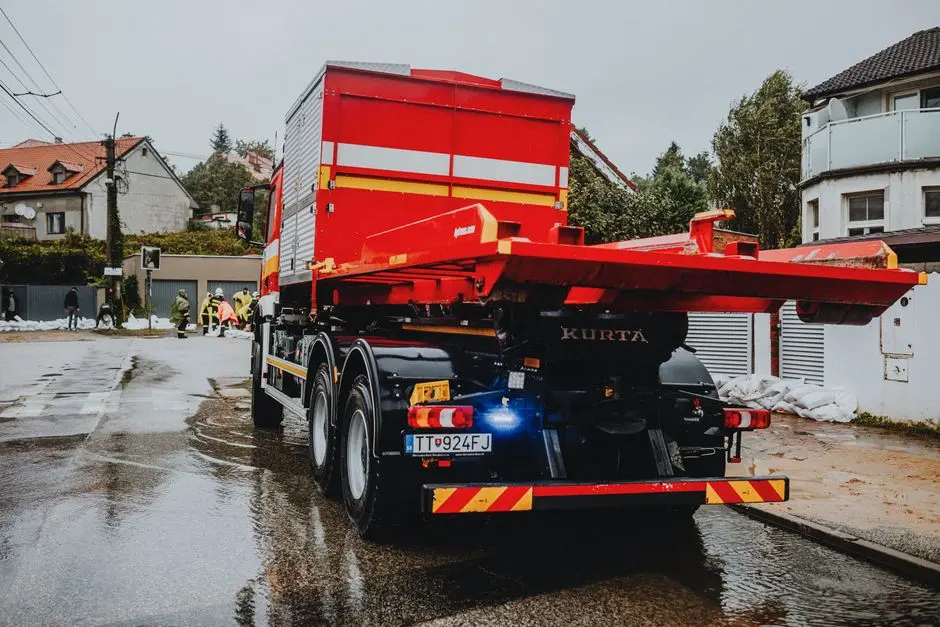 A red tow truck parked on a wet street in an urban neighborhood with workers in the background.