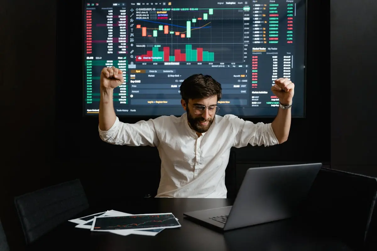 Businessman celebrates stock market success with hands raised in excitement at a trading desk. cfo