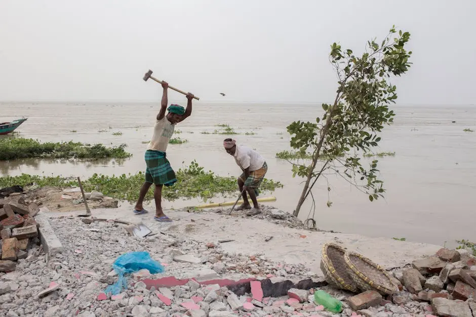 Two men clearing debris by a riverside, using hammers amidst construction materials.