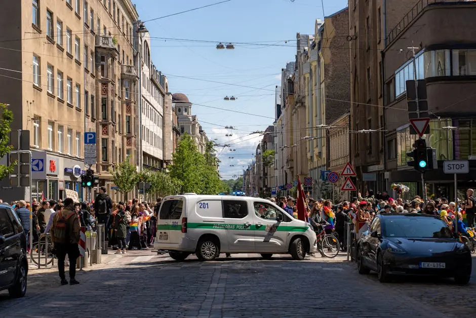 Colorful pride parade with a crowd and police presence in Riga, showcasing urban architecture.