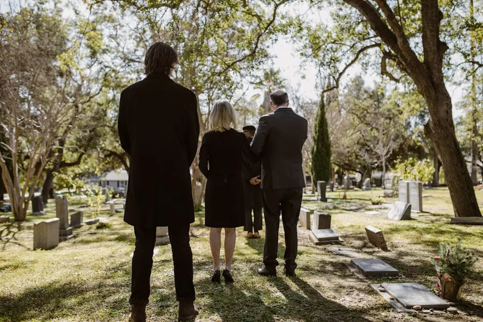 A solemn gathering of adults in formal attire at a serene cemetery setting, beneath trees.