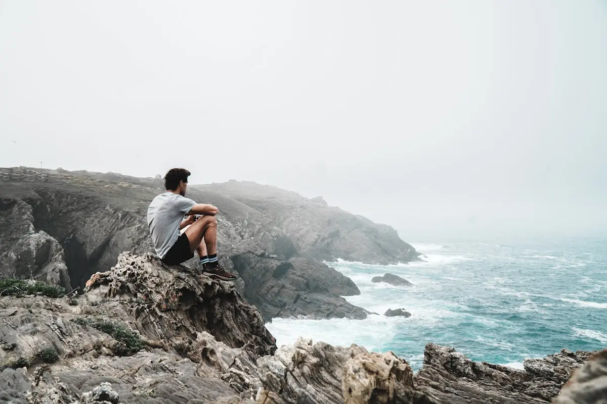 A man sitting on a rocky cliff overlooking a misty ocean coast, contemplating nature.