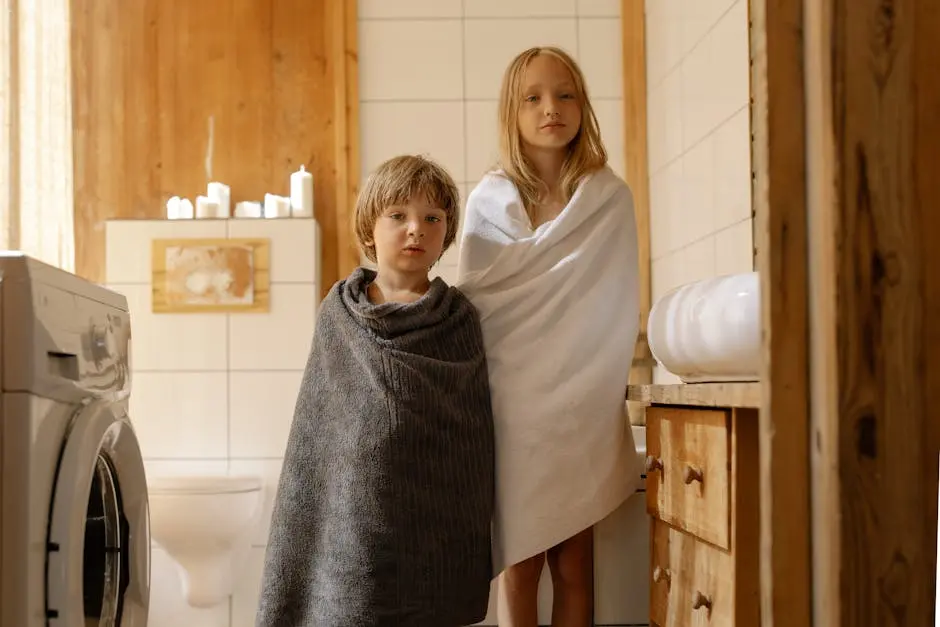 Two children stand wrapped in towels in a warmly lit, wooden bathroom with a washing machine.