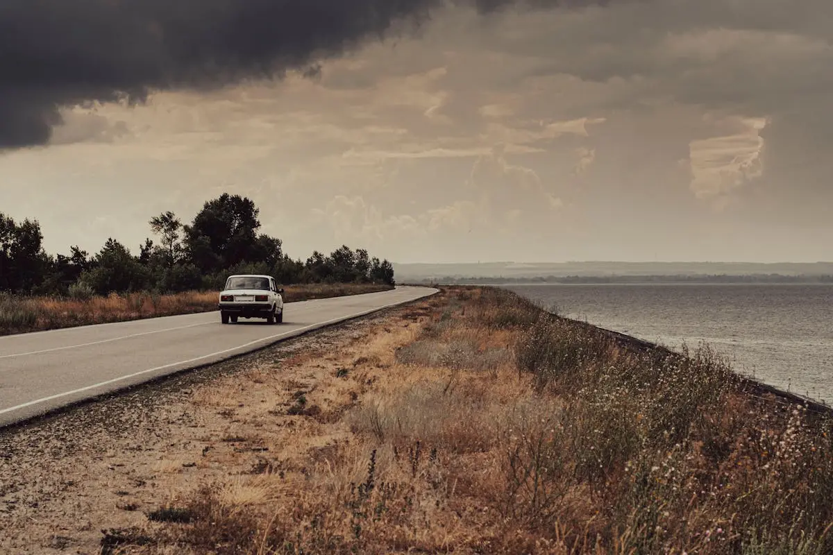 A vintage car travels down a deserted coastal road under dramatic skies at twilight.