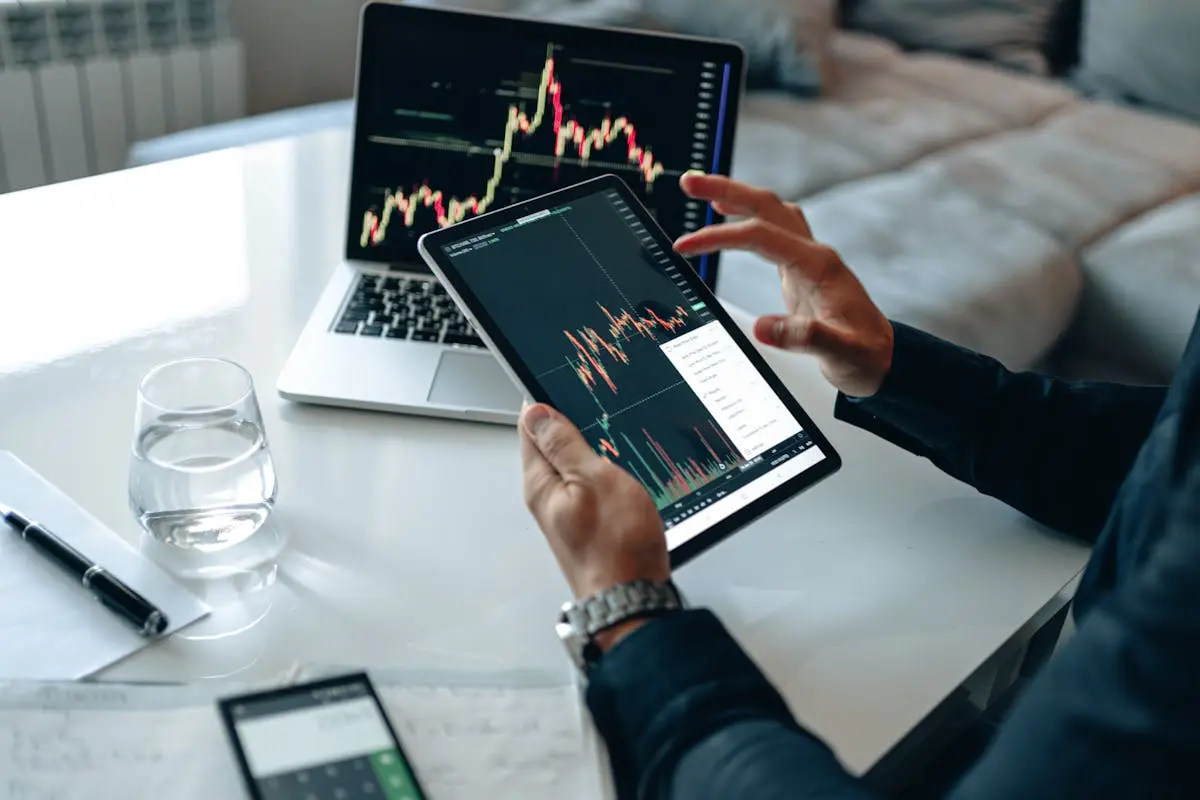 Close-up of a person using a tablet for online trading analysis with a laptop and glass of water nearby.