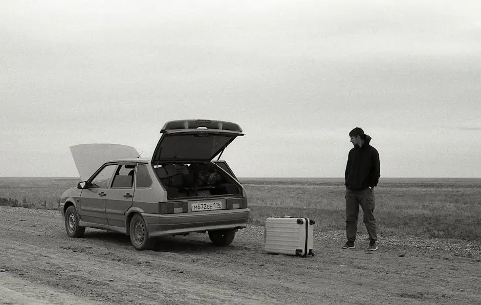 A man stands by a broken down car on a dirt road with luggage in a desolate, rural setting.