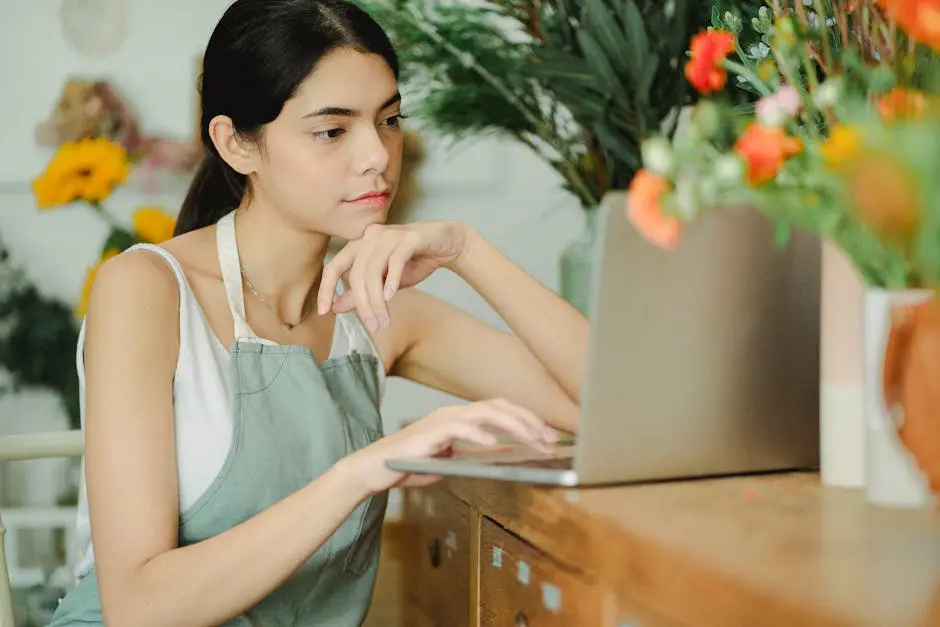 A female florist wearing an apron works on a laptop among colorful flowers in a flower shop.