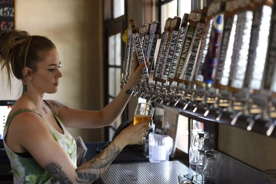 A woman pouring a glass of craft beer in a taproom, showcasing tattoos and a casual atmosphere.