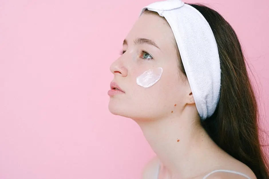 A serene woman applying skincare cream on her cheek against a pink background.