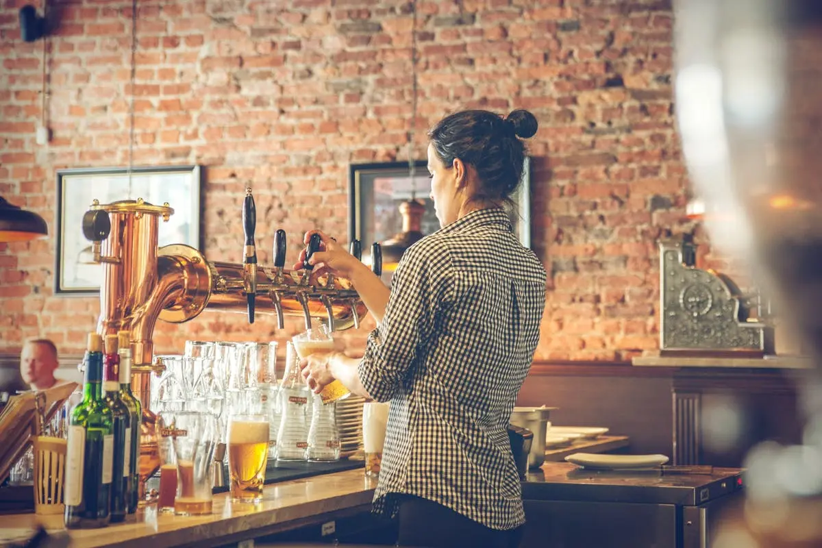 A woman bartender pours beer from taps in a cozy brick-walled bar.