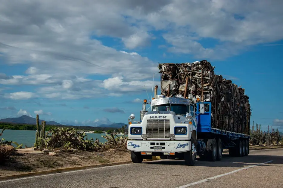 White Mack truck loaded with cargo travels on a scenic road by a lake under a blue sky.