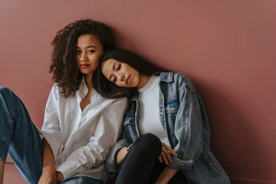 Two women in casual denim and white outfits sitting against a plain wall.