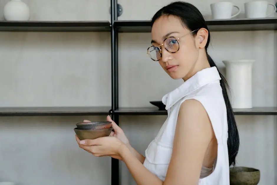 A fashionable woman in glasses holds ceramic bowls against a modern indoor backdrop.
