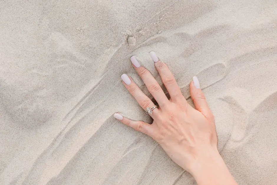 A close-up of a female hand with a diamond ring and manicured nails on sand.