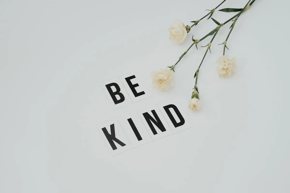 Minimalist image of ‘Be Kind’ text with white flowers on a light background, promoting kindness.