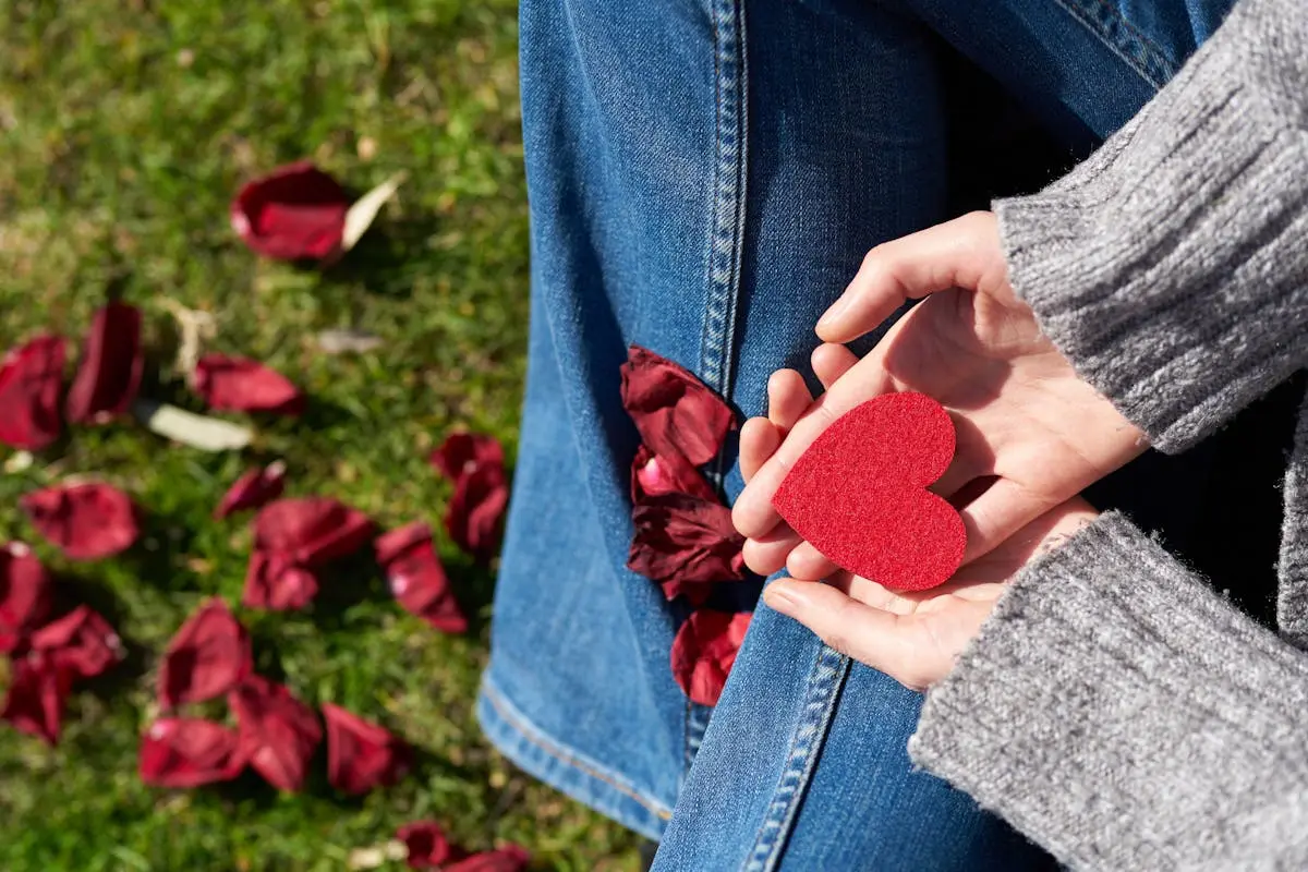 A woman sits on grass holding a red heart-shaped card, surrounded by rose petals, symbolizing love and compassion.