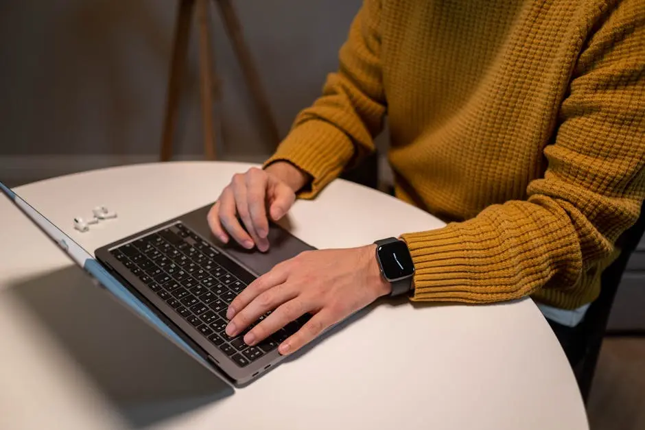 A man in a yellow sweater uses a laptop and smartwatch at a table. AirPods are visible beside him.