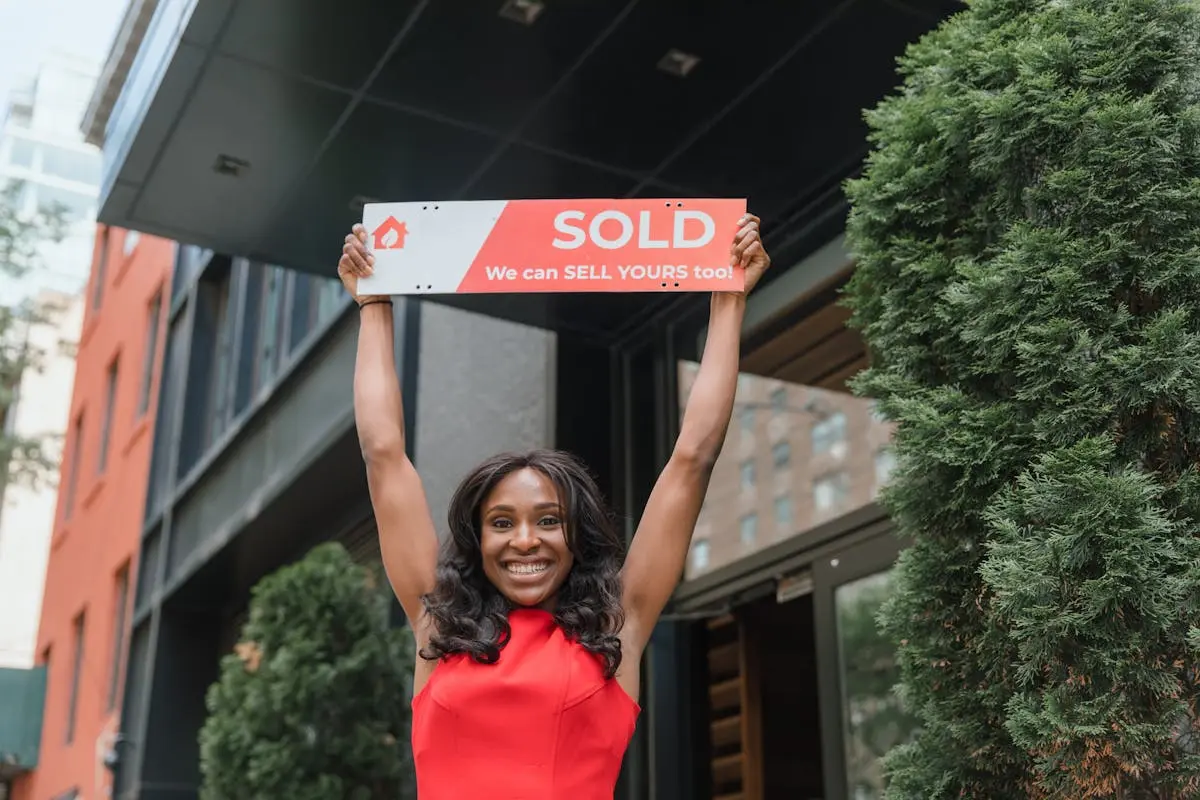 Smiling woman holding a &lsquo;Sold&rsquo; sign outside a building, celebrating a successful sale.