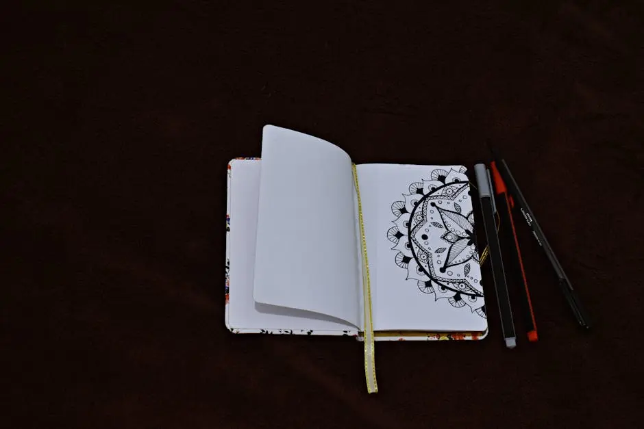 Open notebook with a black mandala drawing, surrounded by pens on a dark background.