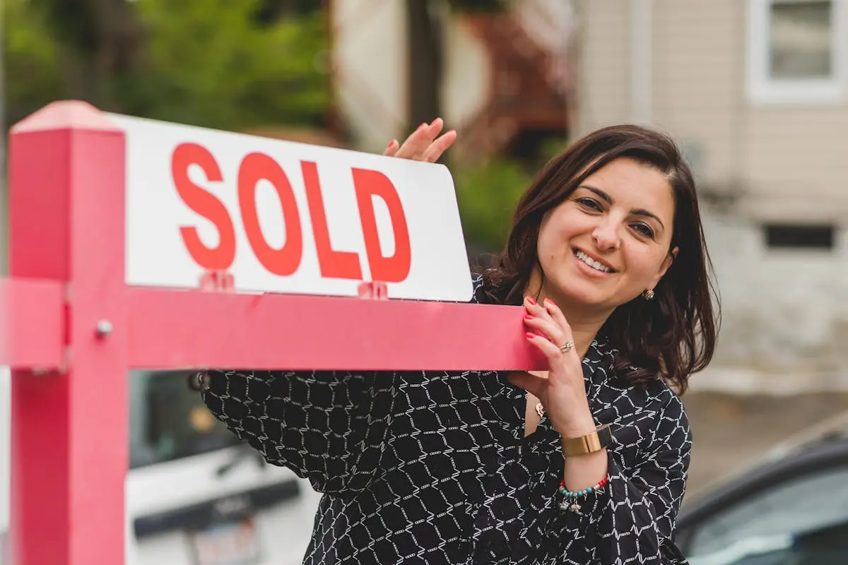 Smiling realtor holding a &lsquo;Sold&rsquo; sign in front of a house. Perfect for real estate marketing.