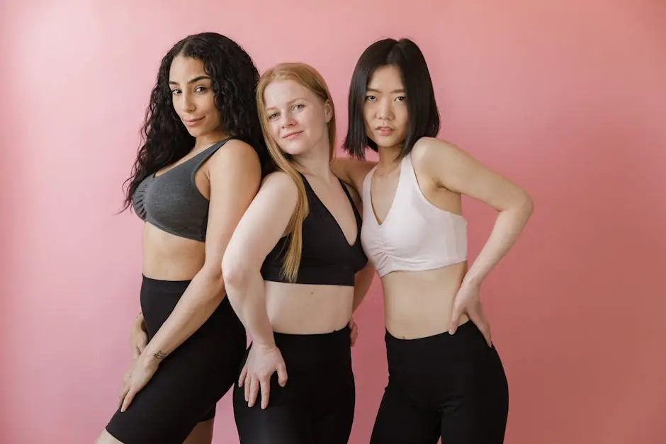 Three women in athletic wear confidently pose against a pink backdrop, showcasing diversity and fitness.