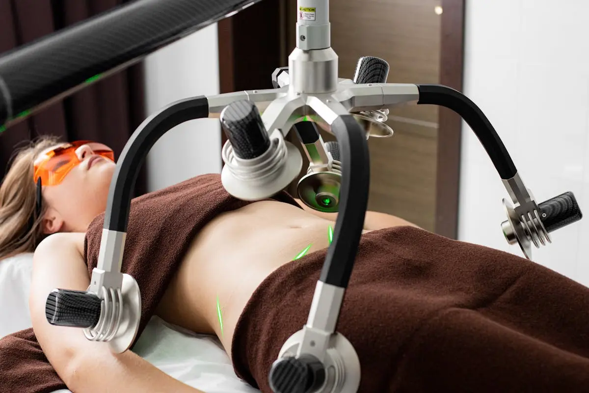 Female client lying on table under lasers of modern weight loss machine on belly during liposuction procedure in medical clinic